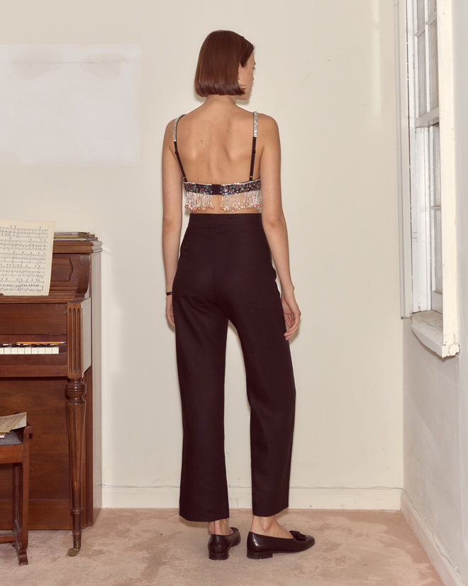A person stands indoors facing away, wearing black pants and the Ritzy Bra near a piano and a window.