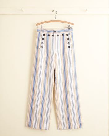 A pair of Riviera Stripe Trousers, crafted from Deadstock ticking fabric with blue, orange, and white vertical stripes and adorned with buttons on both sides, hanging on a wooden hanger against a light-colored wall.