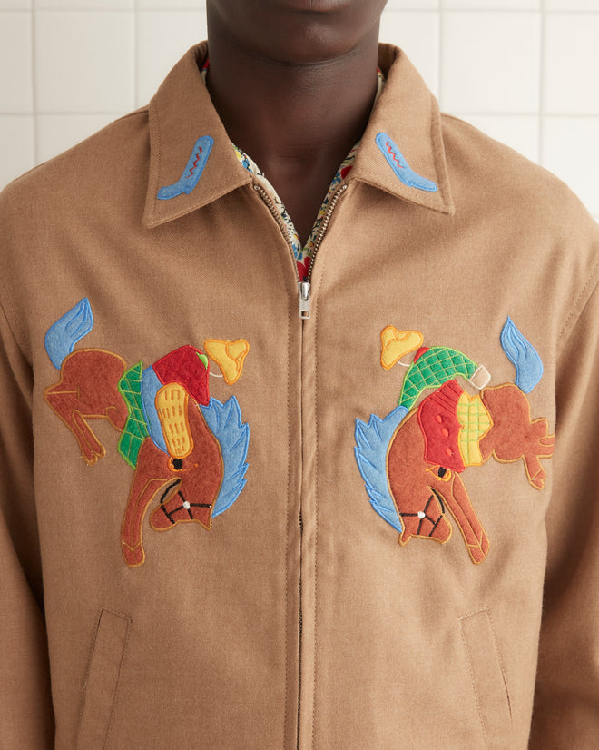 A man stands in a tiled room wearing the Rodeo Ohio Jacket over a patterned shirt, blue jeans with decorative cuffs, and brown shoes.