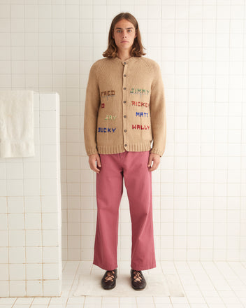 A person with long hair stands in a tiled room, wearing the Roll Call Cardigan, a historical reproduction knitwear piece adorned with multicolored text, paired with pink pants.