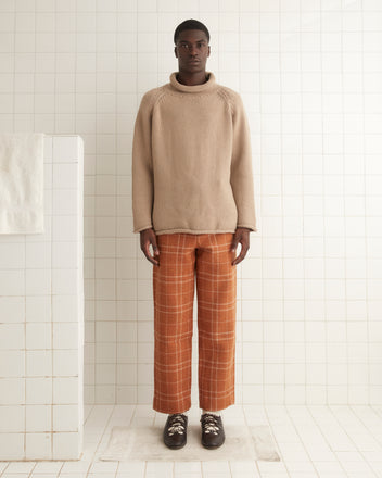 A person standing on a tiled floor, wearing an oversized Cashmere Rollneck - Oatmeal sweater, orange checkered pants, and brown shoes against a white tiled background. The ensemble exudes a 1960s-inspired vibe.