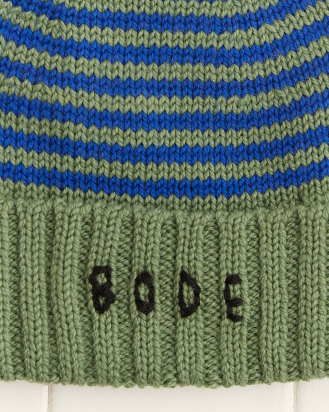 The Roll-Up Tassel Beanie in green is a vintage-style striped beanie made from soft merino wool, highlighted with a black tassel and an embroidered 