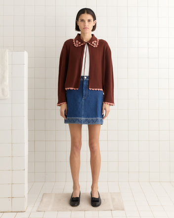 A woman in a Rookie Cardigan adorned with pearl-beaded flowers and a denim skirt stands in a tiled room, looking forward with her hands by her sides.