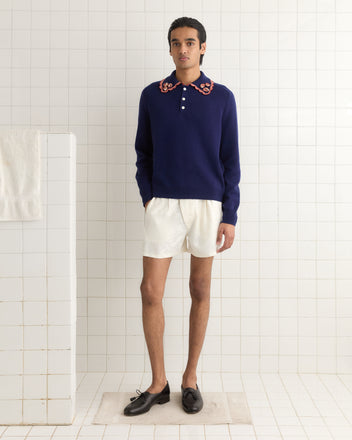 A person in a Rookie Polo with candy-striped trim and white shorts stands in a tiled room wearing black loafers. A towel hangs on the wall rack nearby.