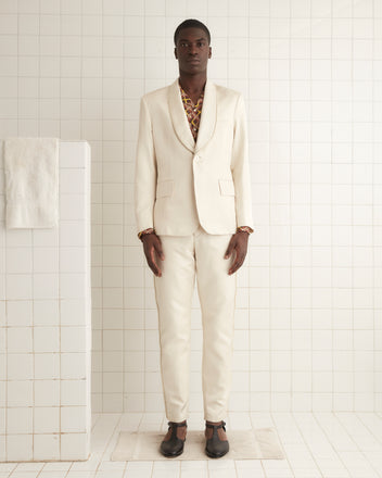 A person in the Roping Shawl Collar Tuxedo Jacket and brown shoes stands against an off-white wall, exuding elegance with its shimmering cream-colored twill fabric accents.