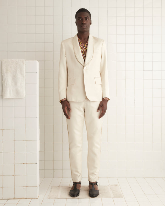 A person in the Roping Shawl Collar Tuxedo Jacket and brown shoes stands against an off-white wall, exuding elegance with its shimmering cream-colored twill fabric accents.