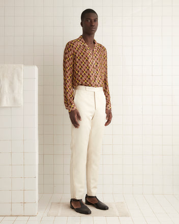 The individual is positioned against a light-colored wall, dressed in an all-white ensemble made from shimmering twill fabric, featuring the Roping Tuxedo Trousers adorned with antique pearl buttons, accessorized with brown sandals and hands tucked into pockets.