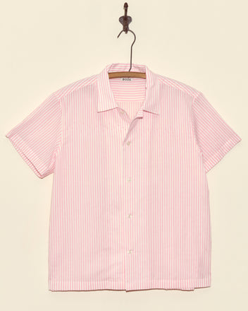 The Rosa Stripe Short Sleeve, a deadstock light pink and white vertical striped button-up shirt, hangs on a hook against a beige wall.