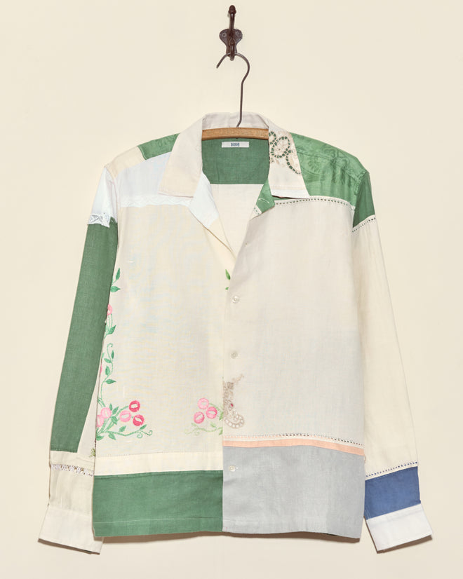 The Rosebud Napkin Shirt - M/L features green, cream, and blue patchwork with floral embroidery and a relaxed collar, shown hanging on a hook.