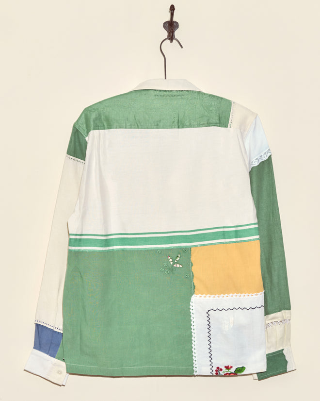The Rosebud Napkin Shirt - M/L, featuring unique green, yellow, white, and blue patchwork panels, hangs on a hook against a plain wall.