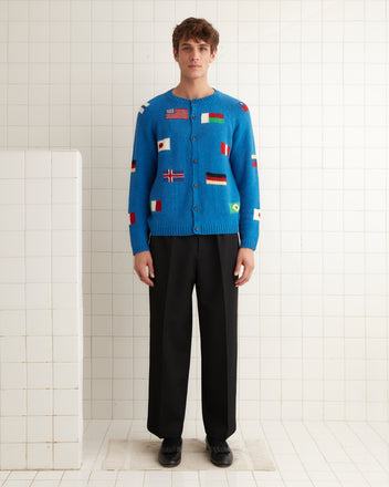 A person wearing a blue Round-the-World Cardigan featuring an intarsia flag pattern, paired with black pants and black shoes, stands against a tiled wall.