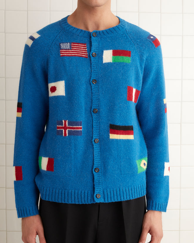 A person wearing a blue Round-the-World Cardigan featuring an intarsia flag pattern, paired with black pants and black shoes, stands against a tiled wall.