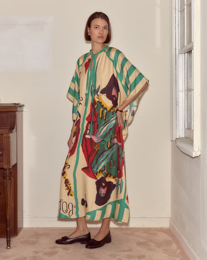 A woman wearing the Royals Kaftan, a long colorful silk garment with green stripes, stands indoors by a window and gazes at the camera.