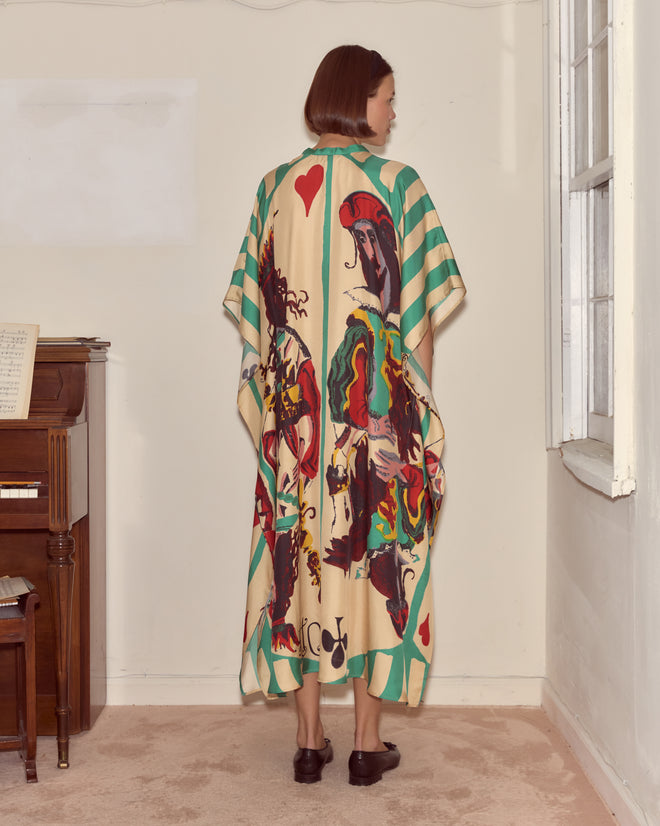 A woman in the Royals Kaftan, featuring a colorful playing-card pattern, stands facing away near a piano and window.