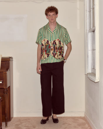 A man in a Royals Short Sleeve Shirt and black pants stands indoors beside a window and a piano.