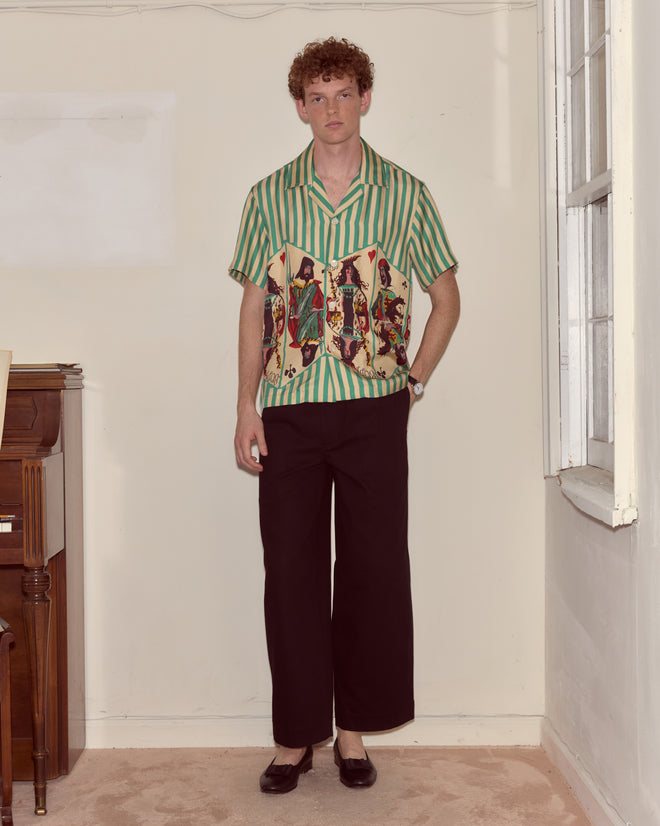 A man in a Royals Short Sleeve Shirt and black pants stands indoors beside a window and a piano.