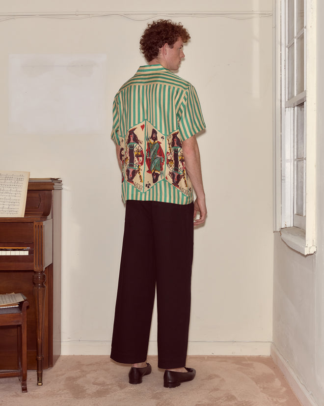 A person in black pants and a vibrant Royals Short Sleeve Shirt stands facing a cream wall near a piano and window.