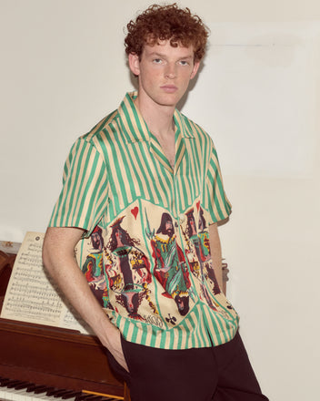 A man wearing the Royals Short Sleeve Shirt stands by a piano with sheet music on it, looking at the camera.