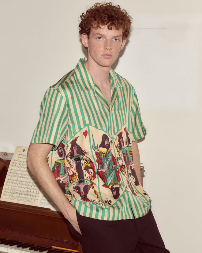 A man wearing the Royals Short Sleeve Shirt stands by a piano with sheet music on it, looking at the camera.