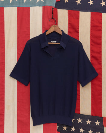The Boxy Polo - Navy, a short-sleeve knit shirt on a hanger, is set against an American flag backdrop and evokes classic 1970s polo style with its timeless cotton knitwear design.
