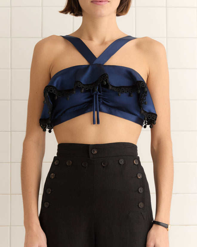 A person wears the Ruffle Bra - Navy, a silky dark blue crop top with ruffles and straps, paired with high-waisted black trousers, enhanced by a backdrop of pristine white tiles.