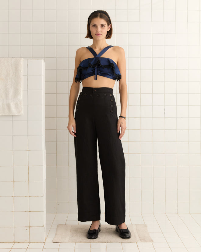 A person in a navy ruffle bra and black high-waisted pants stands in a tiled room, exuding 1950s-inspired elegance.