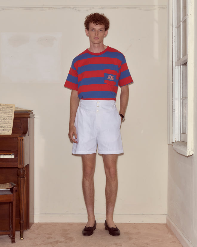 A man in a red and blue striped Clubhouse Tee and white shorts stands indoors by a piano and window, his Bode monogram subtly visible.