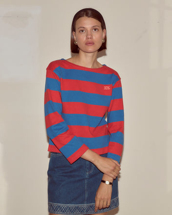 A woman in the Clubhouse Long Sleeve—crafted from 100% cotton with bold red and blue stripes—pairs it with a denim skirt against a plain light wall, creating a classic, comfortable style.