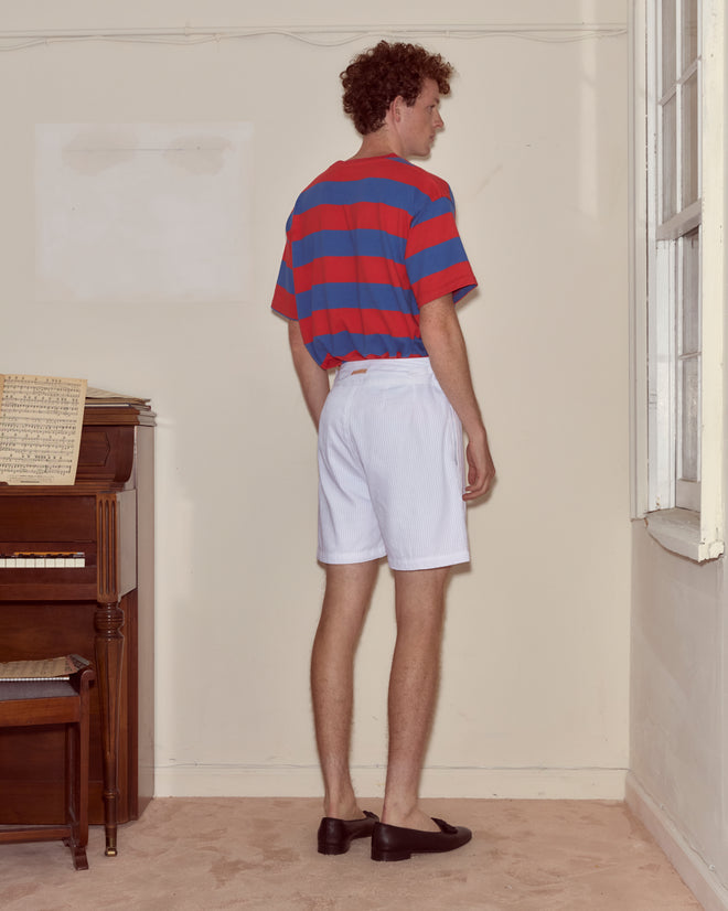 A man in a Bode Clubhouse Tee and white shorts stands near a piano, facing away from the camera.