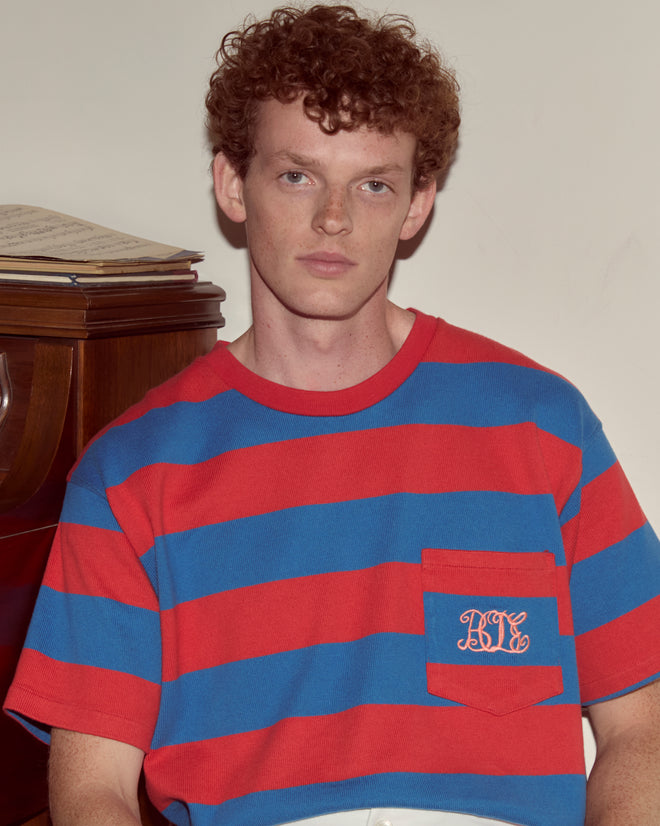 A young man with curly red hair wears the Clubhouse Tee, sitting by a piano with sheet music.