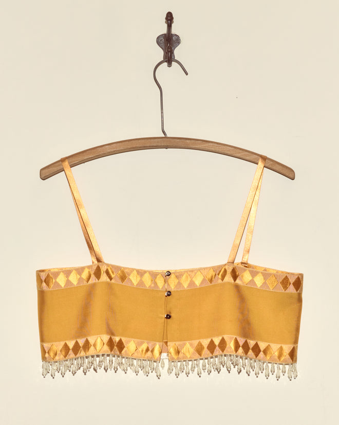 The Saffron Pierrot Bra in size S, featuring thin straps and beaded fringe, crafted from French couture textile, hangs on a wooden hanger against a beige wall.
