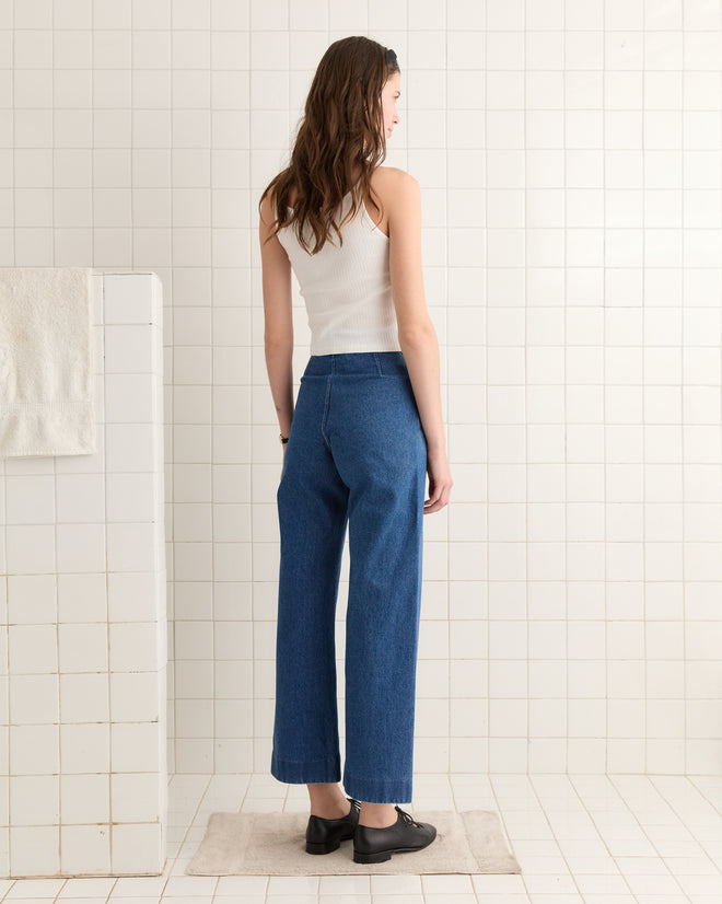 A woman in a Beaded Sailfish Tank and blue jeans stands with her back to the camera in a tiled bathroom.