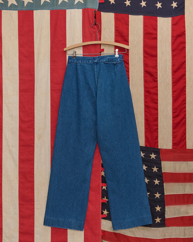 Denim Sailor Trousers crafted from 100% cotton hang on a wooden hanger before an American flag backdrop.