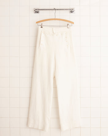 The Linen Sailor Trousers in white, hanging gracefully against a tiled wall, evoke timeless elegance and vintage charm from the 1940s.