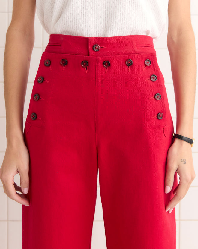 A person wearing the red Spring Twill Sailor Trousers, inspired by 1940s silhouettes and crafted from heavyweight cotton twill with multiple buttons, paired with a white textured top.