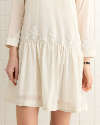 Standing gracefully against a tiled background, the person wears the Salt Marsh Dress, featuring floral embroidery and intricate geometric white beadwork, reminiscent of an antique beaded tunic's elegance.
