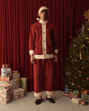 Dressed in the festive Santa Jacket, a person stands beside a beautifully adorned Christmas tree and an assortment of gifts, all framed by a vibrant red curtain that highlights the joyous holiday fashion.