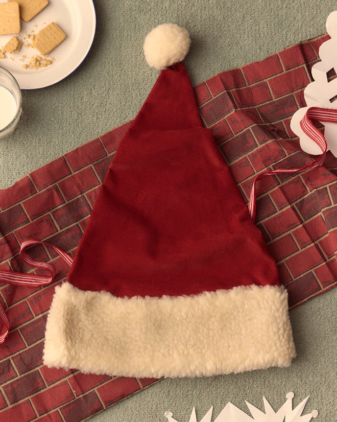 The Santa Hat, featuring red velvet with white trim and a pom-pom, rests on a festive cloth alongside cookies, milk, and snowflake cutouts, embodying classic holiday styles.