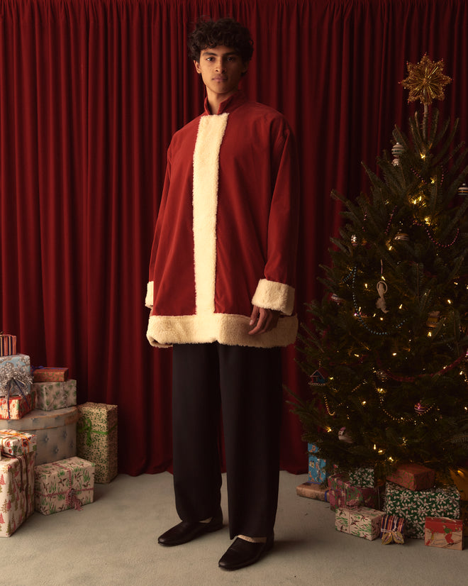 Dressed in a Santa Tunic, a person stands beside a Christmas tree adorned with wrapped gifts, framed by elegant red velvet curtains.