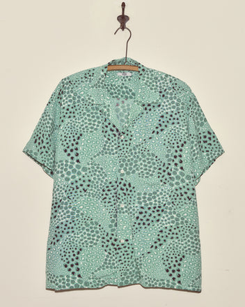 The Sardine School Shirt - XXL, a light green short-sleeve vintage button-up with a circular dot pattern, hangs on a wall hook.
