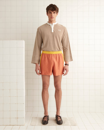 A person wearing a beige long-sleeve shirt and Satin Field Shorts in orange with a contrasting waistband stands in a tiled room.