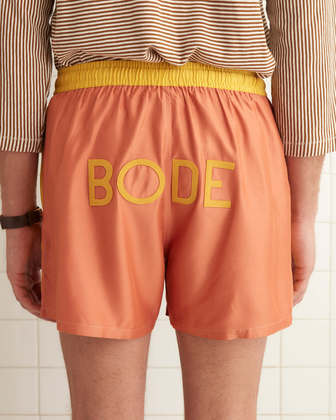 A person wearing a beige long-sleeve shirt and Satin Field Shorts in orange with a contrasting waistband stands in a tiled room.