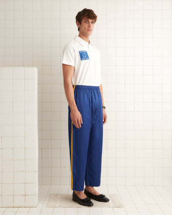 A man in a white polo and Satin Track Pants - Blue stands in the tiled room.