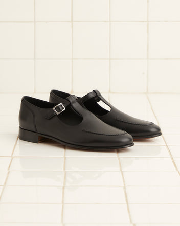 A pair of School Shoes - Black, crafted in glossy black leather with monk strap detailing and made in Italy, rests elegantly on a white tiled floor.