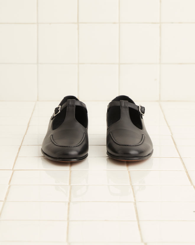 A pair of School Shoes - Black, crafted in glossy black leather with monk strap detailing and made in Italy, rests elegantly on a white tiled floor.