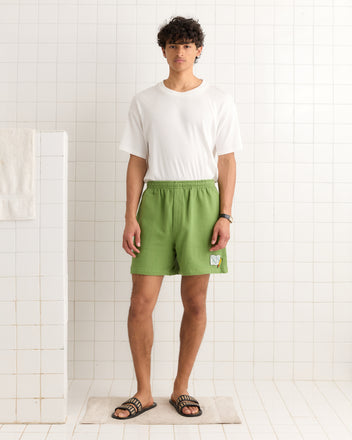 A man in a white t-shirt and green Score Card Shorts stands in a tiled room, wearing black sandals.