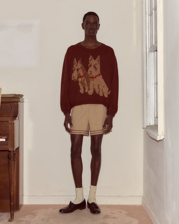 Wearing the Scottie Pullover— a hand-embroidered wool sweater with Scottie dogs— along with beige shorts, socks, and loafers, a person stands indoors by a piano and window.