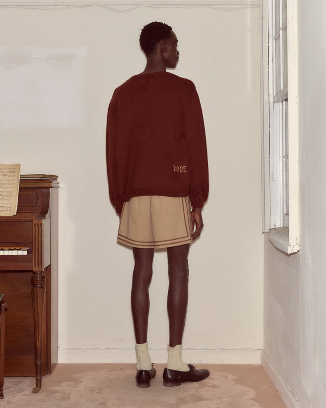 A person wearing the Scottie Pullover, a hand-embroidered wool sweater with Scottie dogs, and beige shorts stands facing a wall near a piano in a softly lit room.