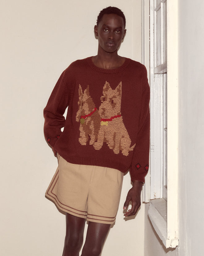 A person wearing the Scottie Pullover—a brown wool sweater with hand-embroidered Scottie dogs—and tan shorts stands by a window in a neutral room.