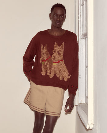A person wearing the Scottie Pullover—a brown wool sweater with hand-embroidered Scottie dogs—and tan shorts stands by a window in a neutral room.